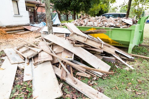 A pile of discarded wooden decking boards and planks is shown on a grassy area adjacent to a construction site in Seven Kings, with a green skip filled with rubble and bricks in the background. The decking materials vary in length and width, featuring weathered, painted, and unfinished surfaces and textures. Some of the wood appears broken or damaged, with rough edges and splintered surfaces. The surrounding environment includes trees, a residential building with a staircase, and a paved pathway, indicating an outdoor location near a property undergoing clearance. The lighting is natural, suggesting daytime, with soft shadows cast across the scene. This image relates to rubbish removal services provided by Office Clearance Seven Kings, highlighting the disposal of construction debris and timber for local property clearance or renovation projects in the area.