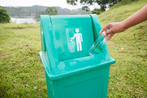 A person disposing of a clear plastic bottle into a bright turquoise wheelie bin marked with a white icon of a person throwing rubbish into a bin, situated outdoors on a grassy area near a body of water and trees in the background. The scene is well-lit with natural daylight, and the bin is positioned on a paved or grassy surface, representing a typical waste disposal setup. The image relates to rubbish collection services provided by Office Clearance Seven Kings, emphasizing environmentally responsible waste disposal in a residential or park setting within the Seven Kings area.
