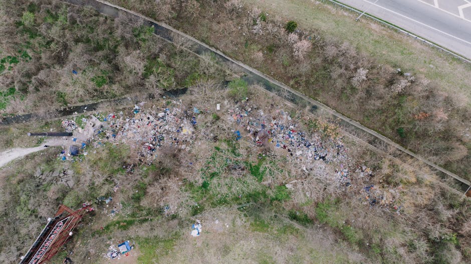 An aerial view of a large, unmanaged debris pile situated on an unpaved, dusty surface next to a gravel and dirt pathway. The mound consists of assorted waste materials, prominently including plastic 