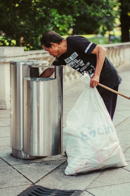On a paved street curb in front of a black metal fence with vertical bars, there are several large, black plastic rubbish bags filled with waste materials. The bags are crumpled and appear to contain
