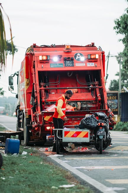 A large accumulation of mixed waste and rubbish bags is gathered around several black, red, and grey recycling bins on a paved parking area, with excess cardboard boxes, paper, plastic containers, and