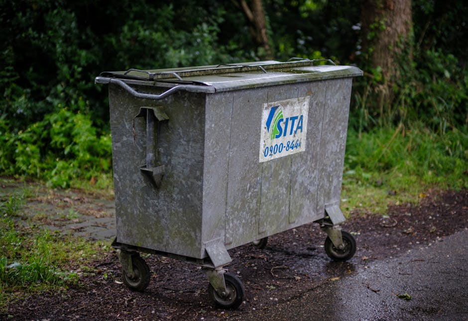 A large, rectangular, grey metal waste container with a hinged lid and four caster wheels is positioned on an outdoor pavement adjacent to a grassy area and trees. The container has a weathered surfac